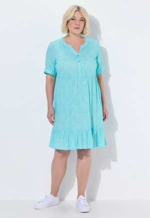 Kvinde står iført en lyseblå knælængde kjole med mønster og knapper samt hvide sneakers mod en ensfarvet hvid baggrund.