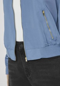 Light blue bomber jacket with a ribbed hem and cuffs, featuring a gold zipper and side pockets, paired with black pants.