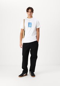 Hvid bomulds t-shirt med blåt grafisk design, sorte afslappede bukser og sorte sko med hvide detaljer, båret med en beige crossbody taske.