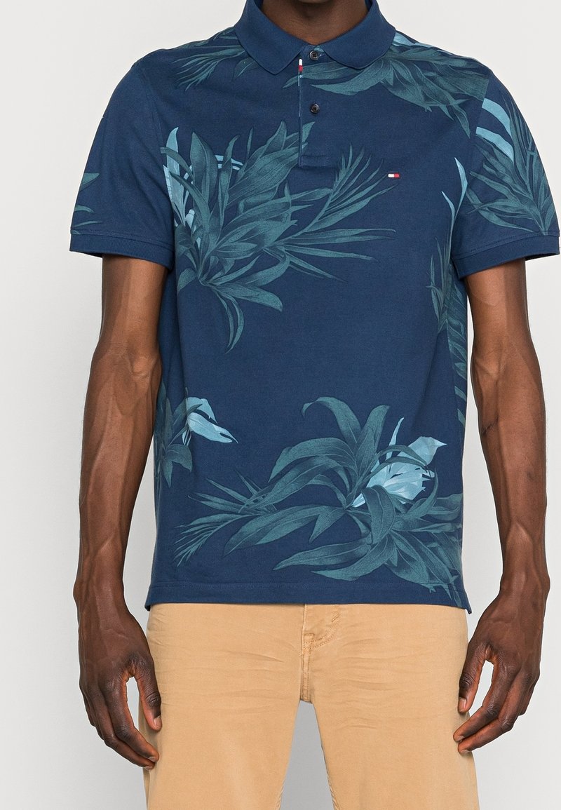 Homme portant un polo bleu marine avec des motifs de feuilles tropicales vertes et un pantalon beige, debout les bras détendus le long du corps.