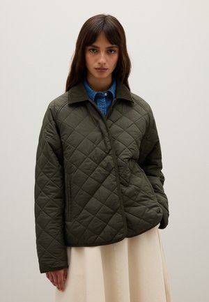 Frau in einer gesteppten olivgrünen Jacke über einem blauen Jeanshemd und einem cremefarbenen Rock, stehend vor einem schlichten hellen Hintergrund.