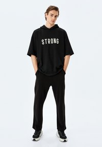 Sudadera negra de gran tamaño con la palabra "FUERTE" en blanco, combinada con pantalones negros y zapatillas deportivas negras con acentos grises.