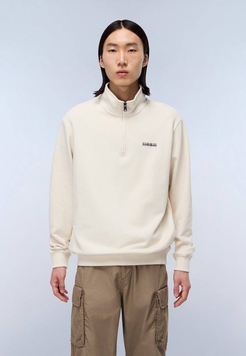 Cream zip-up sweatshirt with a high collar, featuring a small black logo on the left chest, made from a soft, textured fabric.