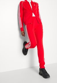 Röd träningsdräkt med vita ränder längs ärmarna och byxorna. Inkluderar zip-up jacka och elastisk midjeband. Bärs med svarta sneakers.