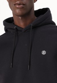 Zwarte hoodie van zachte stof met een voorste kangoeroe-pocket, verstelbare trekkoorden en een klein wit logo op de linkerborst.