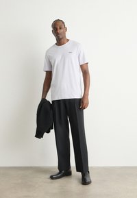 Světle šedé tričko, krátké rukávy, slim fit, s malým logem; kombinováno s černými kalhotami širokého střihu a černou obuví, držící černý svetr.
