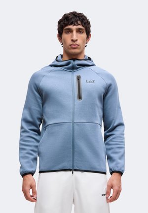 Homme portant un sweat à capuche bleu clair zippé avec poche zippée sur la poitrine, associé à un pantalon blanc sur fond uni.