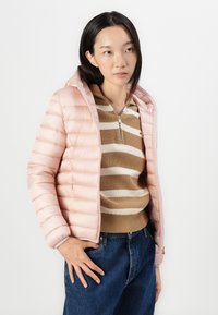 Lys rosa puffer-jakke med horisontal søm, glidelås foran og hette, iført over en brun stripete genser og blå jeans.
