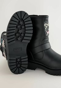Botine negre din piele ecologică, cu un sole din cauciuc texturat cu modele de stele. Accente decorative includ desene florale colorate și fundițe.