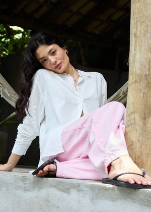 Sieviete ar gariem tumšiem matiem, baltu kreklu un rozā biksēm, sēžot uz betona apmales, atslābusi poza, blakus koka stabam un šūpuļtīklam.