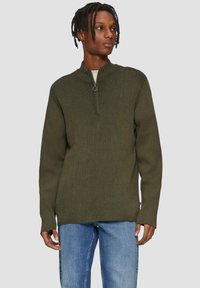 Olivgrüner Zip-Pullover mit gerippter Struktur, langen Ärmeln und einem runden Reißverschluss-Zug, kombiniert mit hellblauen Jeans.