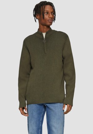 Pull à col zippé vert olive avec texture côtelée, manches longues et tirette circulaire, associé à un jean bleu clair.