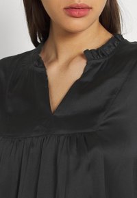 Black satin top with a ruffled neckline and pleated design, featuring a subtle sheen and a V-shaped cutout at the collar.