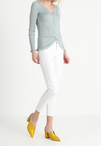 Top a maniche lunghe ribbed di colore azzurro con scollatura a onde e chiusura con bottoni, abbinato a pantaloni bianchi slim-fit e mule gialli.
