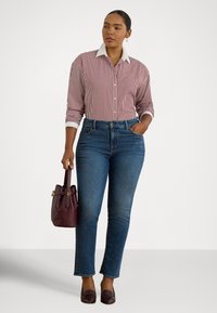 Γυναίκα που φοράει κόκκινο και λευκό ριγωτό πουκάμισο, μπλε τζιν και μπορντό παπούτσια, κρατώντας μια μπορντό τσάντα που ταιριάζει, στέκεται μπροστά σε λευκό φόντο.