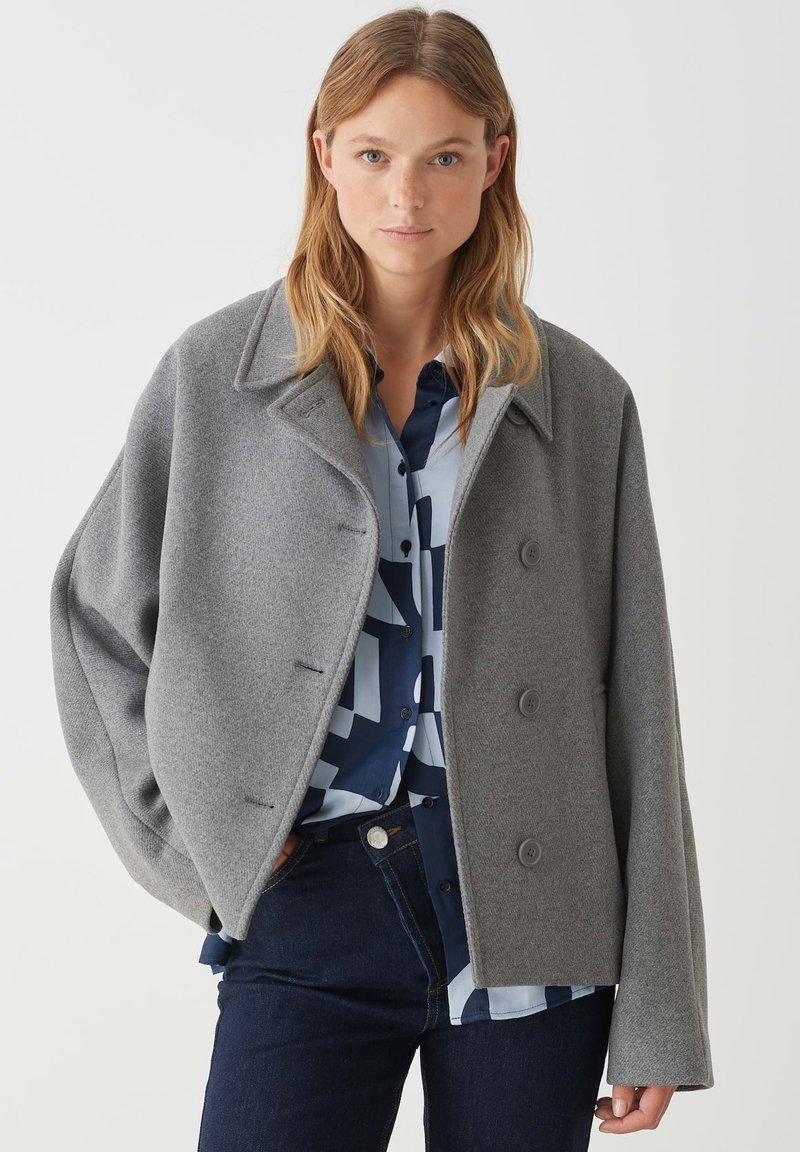 Giacca in misto lana grigia con design doppiopetto, grandi revers e vestibilità ampia. Indossata sopra una camicia blu a fantasia.