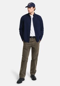 Camicia navy con bottoni, t-shirt bianca, pantaloni verde oliva, cintura nera, stivali neri e berretto da baseball navy con accenti rossi.