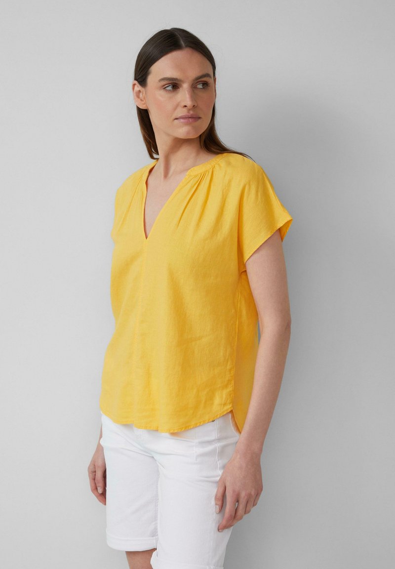 Blouse en lin jaune à manches courtes et décolleté en V avec des détails froncés, dotée d'une fente sur les côtés et d'une coupe décontractée. Portée avec un short blanc.
