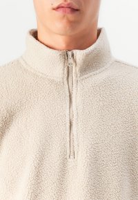 Beige fleece halvhøj zip trøje med en tekstureret overflade, høj krave og sølv lynlås lukning ved halsen. Blødt, varmt materiale.