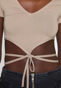 Top corto beige con mangas cortas y un escote en V profundo. Presenta cortes diagonales y lazos ajustables en la cintura. Tejido suave y elástico.