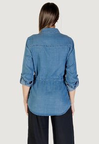 Camicia di chambray azzurro chiaro con maniche lunghe, polsini arrotolati e vita fascata; presenta un collo classico e un orlo curvo.