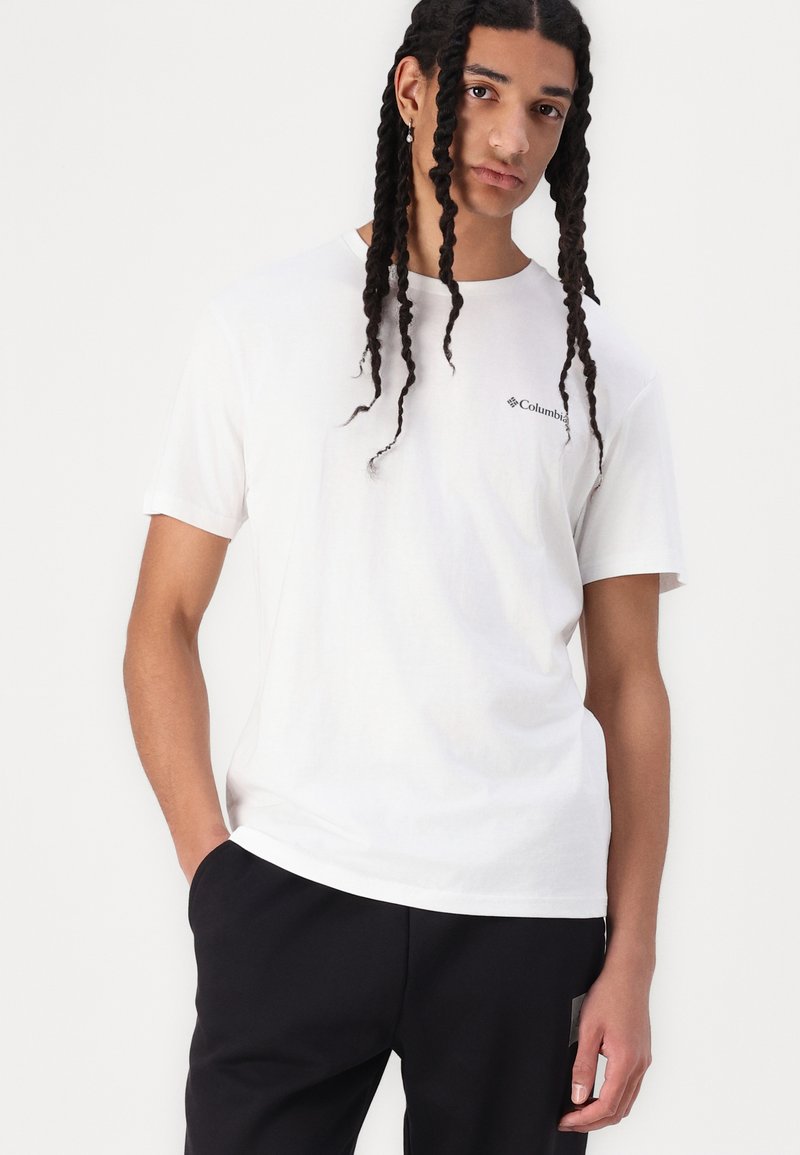 T-shirt en coton blanc avec un col rond et des manches courtes, arborant un petit logo noir de Columbia sur la zone de la poitrine gauche.