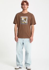 Brun bomulds T-shirt med grafisk tryk i sort, gul og hvid, parret med lyseblå baggy jeans og sort-hvide sneakers.