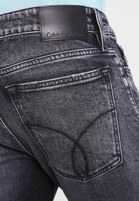 Vaqueros negros de denim con un acabado texturizado, con un parche de logo característico en la cintura y los bolsillos traseros en costuras contrastantes.