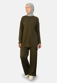 Olive green two-piece outfit with a long top and wide-leg pants. Soft fabric, textured surface, and a relaxed fit. White sneakers complete the look.