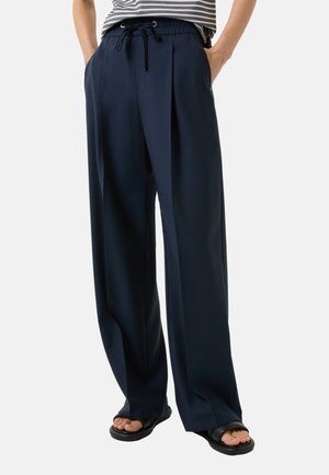 Marineblauwe, wijde broek met een trekkoord in de taille, voorplooien, zijzakken en een soepele textuur. Draagt met zwarte sandalen.