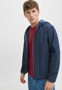 Marineblauer Windbreaker mit Kapuze, weißem Reißverschluss und blauer Innenseite, kombiniert mit einem kastanienbraunen T-Shirt und blauen Jeans.