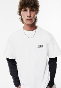 Hvid bomulds t-shirt med rund hals, der har et lille grafisk motiv på venstre bryst og er lagdelt med en sort langærmet skjorte under.