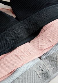 Trois brassières de sport en tissu extensible en noir, rose clair et gris clair, avec une texture côtelée et un logo "NEXT" en relief sur les bandes.