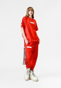 Camiseta roja de gran tamaño con un bolsillo horizontal y pantalones rojos de corte holgado con detalles de flecos y acentos plateados, combinados con botas blancas.