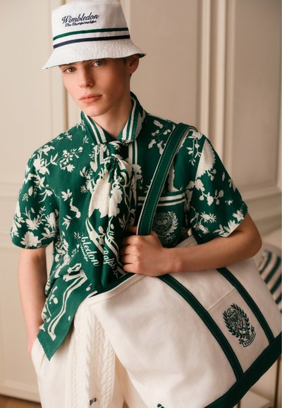 Jeune personne portant un bob blanc Wimbledon, une chemise fleurie verte avec une écharpe, un pantalon blanc et tenant un grand sac fourre-tout blanc avec une bordure verte.