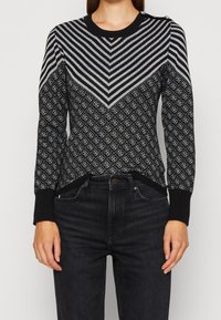 Schwarzer Strickpullover mit einem Chevron-Streifenmuster in metallic Silber und einem geometrischen Design. Mit gerippten Bündchen und Ausschnitt sowie Knopfakzenten.