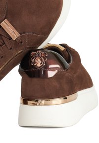 Brown suede sneaker with a white sole, featuring a glossy brown heel cap adorned with a decorative emblem and gold accents.
