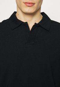 Polo noir avec un col classique et un tissu texturé. Le design comprend une patte de boutonnage à trois boutons et une coupe légèrement ajustée.