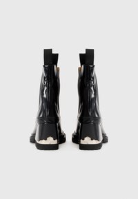 Une paire de bottines noires en cuir avec des languettes et une accent métallique argenté sur les talons épais, présentée de dos sur fond blanc.