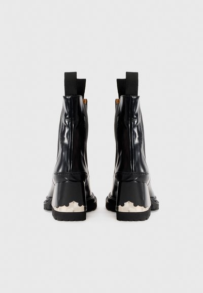 Une paire de bottines noires en cuir avec des languettes et une accent métallique argenté sur les talons épais, présentée de dos sur fond blanc.
