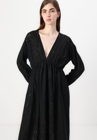 Μαύρο φόρεμα με βαθύ V ντεκολτέ, μακριά μανίκια και μαζεμένη μέση. Ο ιστός έχει μια υφή με λουλούδια, προσφέροντας μια ρέουσα σιλουέτα.