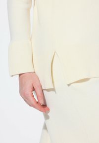 Kremowy sweter dzianinowy z prążkowanymi mankietami, z bocznymi rozcięciami dla większej swobody ruchów. Miękka tekstura i prosty design podkreślają jego minimalizm.