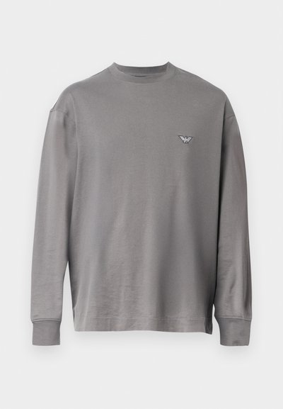 Pelēks garām piedurknēm t-krekls, izgatavots no mīkstas auduma, ar apaļu kakla izgriezumu un mazu izšuvumu uz krūtīm. Kopumā tam ir gluda tekstūra.