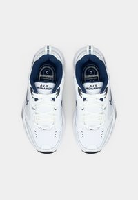 Sapatilhas brancas Nike Air Monarch com interior azul-marinho, apresentando um design com atacadores, detalhes perfurados e acentos de logotipo na língua.