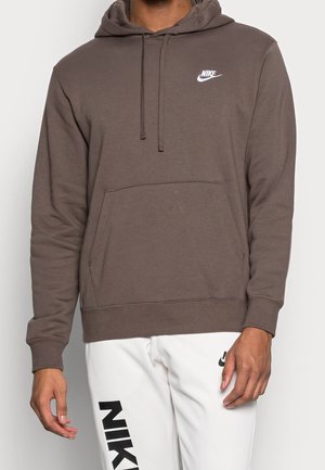 Muž nosící hnědou mikinu Nike s přední kapsou a bílé kalhoty Nike s černým logem na levé noze, stojící proti jednoduchému pozadí.