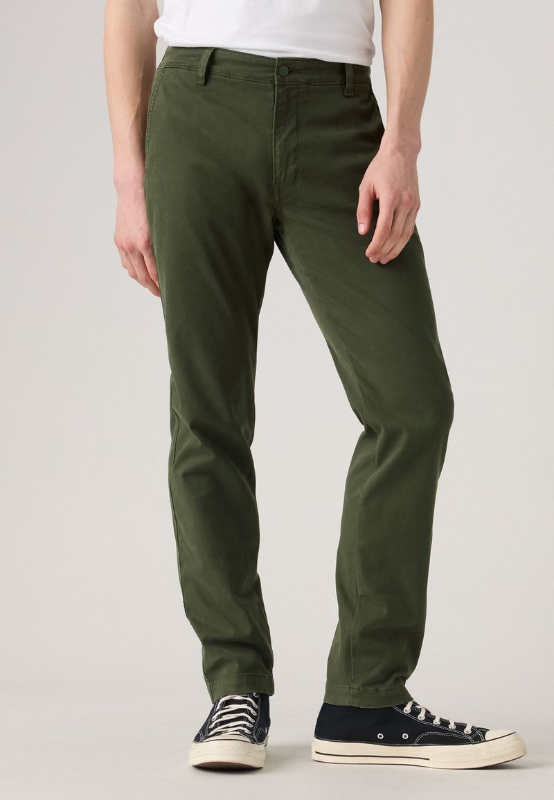 Lower body of person wearing olive green pants and black high-top sneakers with white soles against plain background.