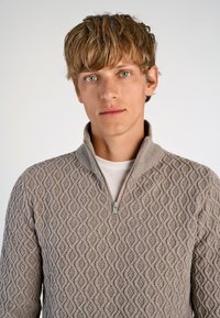 Lichtbruine, gestructureerde trui met een ruitpatroon, voorzien van een ritssluiting bij de kraag en met lange mouwen, gedragen over een wit shirt.