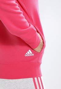 Rosa hoodie i mjukt material med en känguruficka, försett med vitt adidas-logotyp och ribbat nederkant. Ärmlängd till armbågen är synlig.