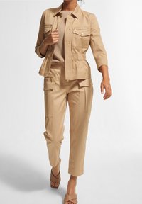 Mujer caminando con una chaqueta de utilidad beige con cremallera y bolsillos de solapa, pantalones cargo a juego, top de punto y sandalias de tacón en color beige.