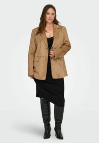 Brauner Wildlederblazer mit Reverskragen, zwei Vordertaschen und lockerer Passform, kombiniert mit einem schwarzen Kleid und kniehohen schwarzen Stiefeln.